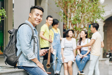 Wall Mural - Portrait of handsome young Vietnamese university student with backpack texting and lookng at camera