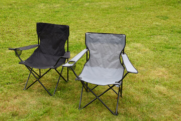 Two grey and black empty folding camping chairs in green lawn grass outdoors in sunny summer day. Camping equipment.
