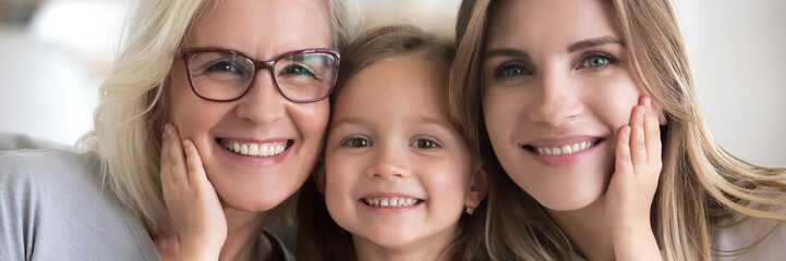 Wall Mural - Little girl her young mother and mature grandma portrait. Multi generational women faces smiling looking at camera close up view photo, family bond concept. Horizontal banner for website header design