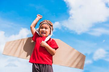 Wall Mural - Happy Asian funny child or kid little boy smile wear pilot hat and goggles play toy cardboard airplane wing flying raises hand up against summer blue sky cloud background, Startup freedom concept