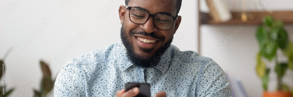 Smiling African businessman sit at workplace holding mobile phone, texting sms, browsing internet. Distant communication, virtual chat usage concept. Horizontal photo banner for website header design - obrazy, fototapety, plakaty 