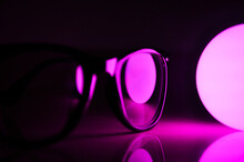 Specs Spectacles Frame Placed Near Light Bulb With Black Background And Colorful Lighting Colours Like Blue Yellow White Purple And Green