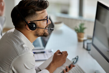 Happy call center agent taking notes while working on desktop PC in the office.