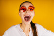 Cheers Cheers !! Girl screams with her mouth wide open and looking through pink glasses on a yellow background