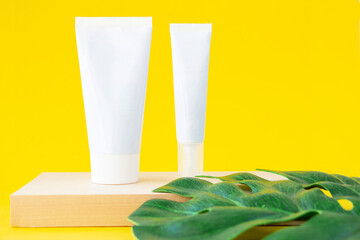 Two empty cosmetic tubes stand on a wooden board, a green leaf of monstera on a yellow background. Copy space for text. Mock branding.