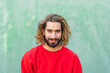 © VISTA by Westend61 - Portrait of bearded young man wearing red sweatshirt in front of green wall