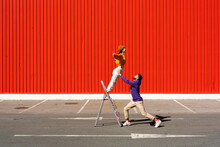 Young Man And Woman Performing With A Ladder In Front Of A Red Wall