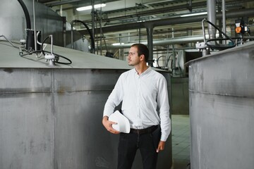 A young handsome employee of a modern factory in a white hard. In the background is a large shop for the production.