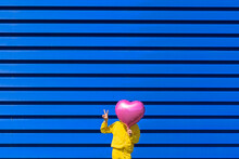 Little Girl Standing In Front Of Blue Background Hiding Behind Pink Balloon And Showing Victory Sign