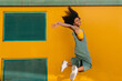 © VISTA by Westend61 - Portrait of happy young woman wearing overalls jumping in front of yellow wall