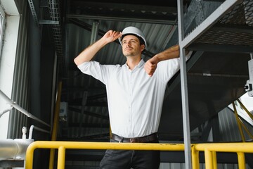 Wall Mural - Young smiling professional in overalls and protective helmet standing in front of camera inside large machinebuilding plant