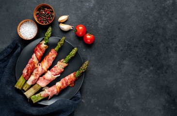 
Baked asparagus with bacon and spices on a black plate on a stone background with copy space for your text