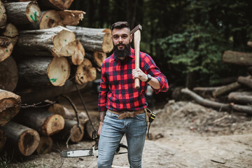 Wall Mural - Young adult lumberjack or logger standing and posing in the woods. He holding big axe and chainsaw.