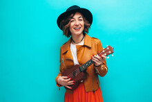 Handsome Young Hipster Girl Wearing Black Leather Jacket And A Hat Plays Ukulele Guitar Isolate Over Blue Background.