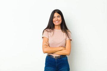 young pretty latin woman looking like a happy, proud and satisfied achiever smiling with arms crossed against flat wall