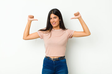 young pretty latin woman feeling proud, arrogant and confident, looking satisfied and successful, pointing to self against flat wall