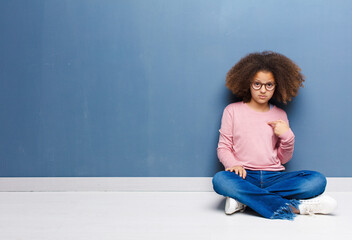 Sticker - african american little girl feeling confused, puzzled and insecure, pointing to self wondering and asking who, me? sitting on the floor