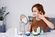 © Eva March - Woman sitting at mirror with bottles of cosmetics products of cream, lotion, mask, spray and scrub, applying face cream with hand. Beauty routine at home, self skincare during isolation.