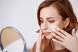 © Eva March - Close up woman looking at mirror applying white mouisturizing face cream with hands. Beauty daily routine at home, self skincare during isolation for glowing healthy skin.