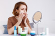 © Eva March - Woman sitting at mirror with bottles of cosmetics products of cream, lotion, mask, spray and scrub, applying face cream with hand. Beauty routine at home, self skincare during isolation.
