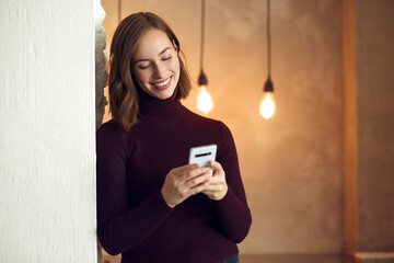 Wall Mural - Portrait of young smiling woman using her phone and looking into it, while standing against a grey wall with warm lightning in the background making a cozy atmosphere.