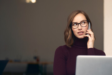 Wall Mural - Beautiful young woman sitting in front of her laptop, talking on her smartphone 