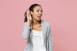 © ViDi Studio - Curious african american business woman in grey suit white shirt isolated on pink background studio. Achievement career wealth business concept. Mock up copy space. Try to hear you with hand near ear.