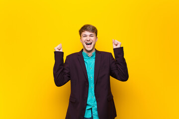 young blonde man looking extremely happy and surprised, celebrating success, shouting and jumping isolated against flat wall