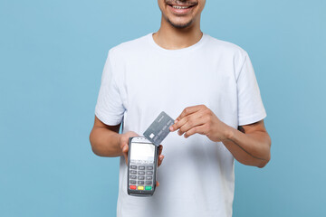 Wall Mural - Cropped image of young african american guy in white t-shirt isolated on blue background. People lifestyle concept. Hold wireless modern bank payment terminal to process acquire credit card payments.