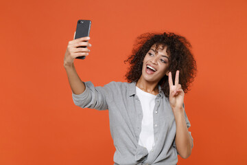 Cheerful young african american woman in gray casual clothes isolated on orange wall background. People lifestyle concept. Mock up copy space. Doing selfie shot on mobile phone, showing victory sign.