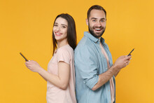 Cheerful Young Couple Friends Guy Girl In Casual Clothes Isolated On Yellow Wall Background. People Lifestyle Concept. Mock Up Copy Space. Standing Back To Back Using Mobile Phone Typing Sms Message.