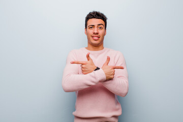 Wall Mural - young arabian man looking puzzled and confused, insecure and pointing in opposite directions with doubts against gray wall