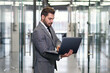 © gorynvd - Smiling bearded business man focused on company datas while standing in office corridor and using laptop.