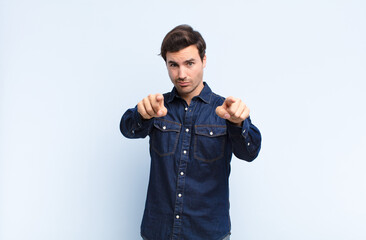 Wall Mural - young handsome man pointing forward at camera with both fingers and angry expression, telling you to do your duty against blue wall