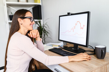 Young woman is using PC for work from home office, woman in eyeglasses looks at computer screen with a graphs on it