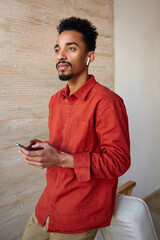 Canvas Print - Portrait of young pensive short haired curly brunette guy with dark skin looking thoughtfully ahead while keeping mobile phone in raised hands, posing over beige interior