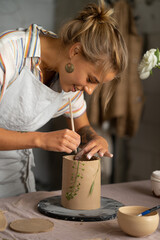 Smiling beautiful girl in blue apron holding rolling pin and happily working with clay at pottery class studio. male potter master rolling up the clay on table with ceramic products.