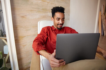 Wall Mural - Positive young pretty short haired dark skinned businessman working out of office with modern laptop, wearing red shirt while sitting in chair over home interior