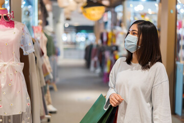 Wall Mural - woman holding shopping bag and looking to clothes at mall and her wearing medical mask for prevention from coronavirus (Covid-19) pandemic. new normal concepts