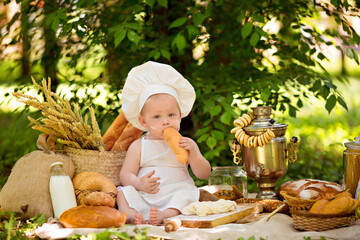 Healthy eating concept. Happy little boy is cooking on nature on a sunny summer day. Toddler baker on a picnic sits making dough and eats bread with milk in a white apron and cap.