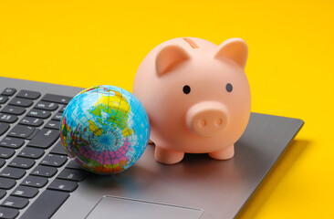 Piggy bank with globe on laptop keyboard,  yellow background.