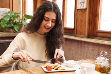 Canvas Print - Image of woman smiling and eating pancakes while sitting in cozy cafe