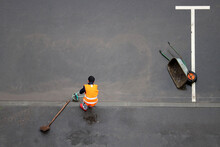 Worker Cleans The Street From Dirt After The Rain. Janitor With Broom, Scoop And Wheelbarrow On The Wet City Road, View From The Top