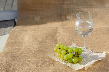 Still life and food photo. Picnic in nature on a sunny day. A bunch of green grape berries, a glass of clear water stand on burlap fabric on an old wooden floor