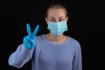 Victory over covid-19. Woman in a medical face mask and gloves shows a victory symbol on a black background.