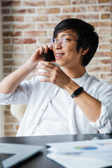 Poster - Image of young asian man using cellphone and drinking coffee in office