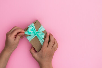 Hands giving or receiving a gift in craft paper with a mint ribbon on pastel pink background. Top view. Copy space.