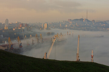 Fog during the day in a commercial sea port in the summer, the tops of port cranes sticking out of the fog, in the background a city on the beach.