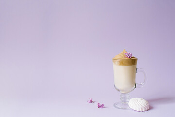 Beautiful dalgona drink a frothy coffee in a transparent mug and flowers lilac to purple Il purple background. Next to the mug is a marshmallow. Copy space