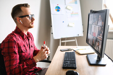 Wall Mural - Online meeting of a young man with his subordinates. He sits accross from the window talking via web camera on PC to a group of people at the workplace, flipchart on the background